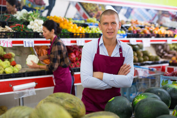 European man in uniform standing among shelves in greengrocer and looking in camera. His female...