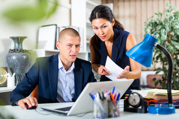 Upset office employe sitting at table and looking on laptop screen on background with disgruntled...