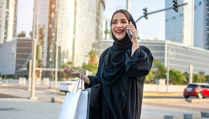 Smiling Arab woman in traditional wear holding shopping bags in hands and talking on mobile phone...
