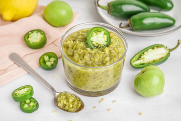 Bowl of tasty green salsa sauce and jalapeno peppers on light background