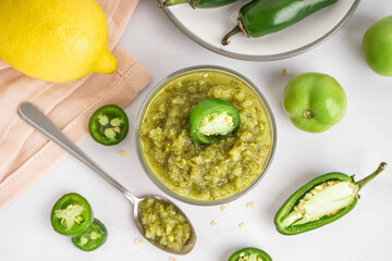 Bowl of tasty green salsa sauce and jalapeno peppers on light background