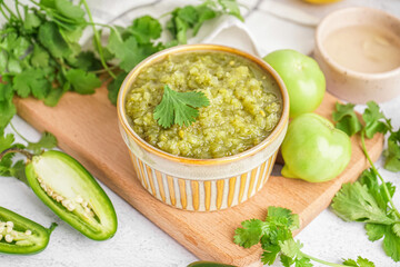 Wooden board with bowl of tasty green salsa sauce, jalapeno peppers, tomatoes and cilantro on table