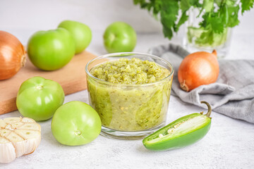 Glass bowl of tasty green salsa sauce and tomatoes on light background