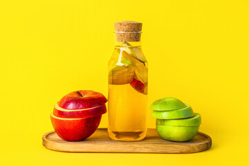 Wooden board with bottle of apple cider vinegar and fruits on yellow background