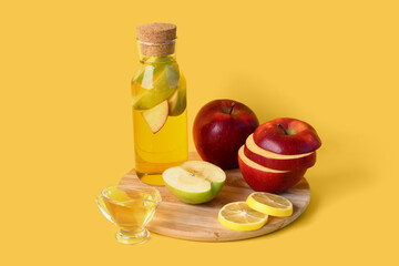 Wooden board with bottle of apple cider vinegar and fresh fruits on yellow background