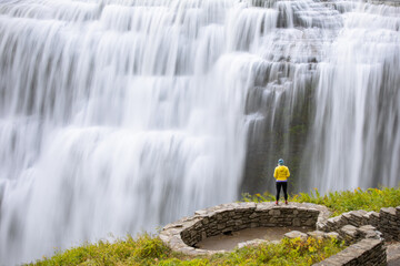 Solo female hiker stands near the edge of a cliff overlooking a giant waterfall. Letchworth State...