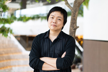 The portrait of Asian man in a black casual shirt with crossed arms standing in front of a blurred background of an office workspace.