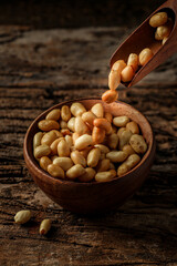 Old and rusty wooden table with a wooden bowl with shelled peanuts and some peanuts falling out of the wooden bowl, selective focus