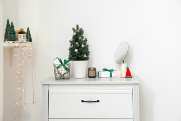 Small Christmas tree with presents, candles and mirror on chest of drawers near light wall