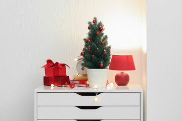 Small Christmas tree, alarm clock, books, presents and glowing lights on chest of drawers near light wall