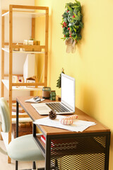 Laptop with Christmas toys and candle on table near yellow wall in office