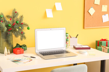 Workplace with laptop, fir branches in vase and Christmas presents near yellow wall