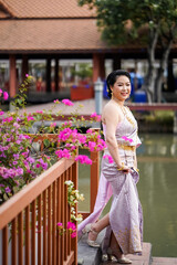 Naklejka premium A woman in a traditional Thai costumes is standing on the wooden bridge while being taken a portrait photo shooting.