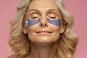 Closeup portrait of beautiful mature woman using under eye patches, doing anti aging procedures isolated on pink background. Skin care, natural beauty concept 
