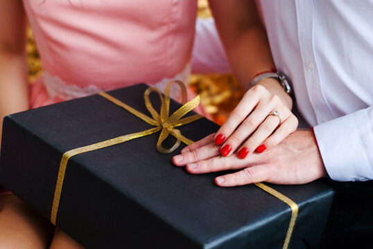Black gift box with golden bow in hands of couple in love. Velvet wrapping paper. Valentine's Day gifts. Couple in love prepares holiday surprises for each other. Ring on female hand with red manicure