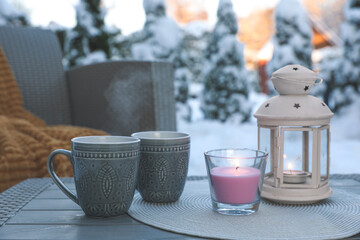Burning candle, lantern and cups with hot drink on coffee table outdoors. Cosy winter