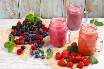 Mason jars of different berry smoothies and fresh ingredients on white wooden table, above view