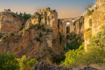 Daytime panoramic image of the clifftop village of Ronda, in Andalucia, Spain, at sunset. The Puente Nuevo bridge and cliffs are bathed in golden light.