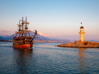 Fototapeta na wymiar Wooden ship and Old Alanya Deniz Feneri lighthouse in Alanya port at sunset. Popular tourist destination in Turkey. Summer travel vacation. View from embankment to beacon.