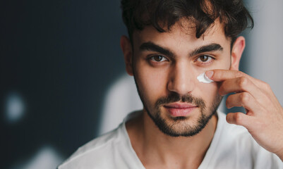 Head shot of handsome young man applying anti-aging moisturiser on face isolated on studio background looking at camera. Concept of fairness and skincare.