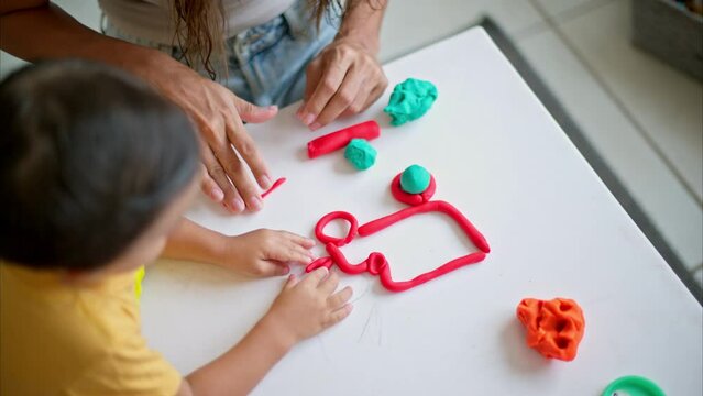 Close up shot of a brunette boy with his mom creating a truck figure out of red and blue play dough on a white table having fun