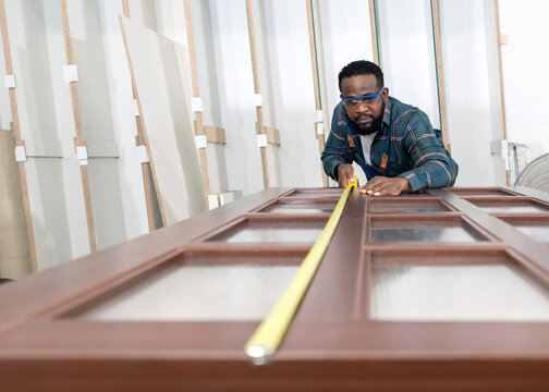 Short Black Hair Man With Moustache And Beard Use A Tape Measure To Measure The Size Of A Wooden Door. Plywood Storage Compartment Are On The Background. Carpenter Worker Work In A Furniture Factory.