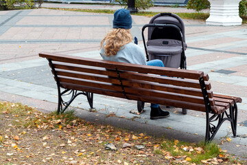A young woman, a mother, is sitting on a bench near a baby carriage with her child