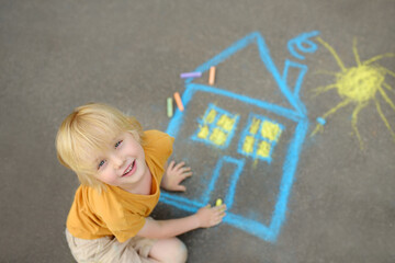 Little child boy is drawing house and sun painted with colored chalk on asphalt of sidewalk. Kids...