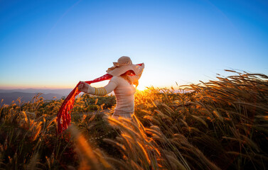 Woman in a hat standing and waving red cloth against the wind with sunset. A young girl stands...
