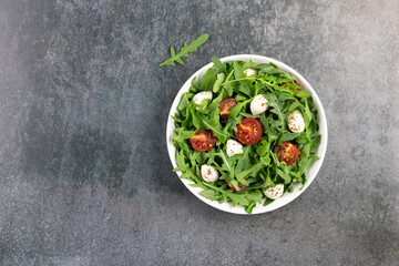 Green salad with arugula, cherry tomatoes, mozzarella cheese, olive oil and spices in a white plate on gray background. 