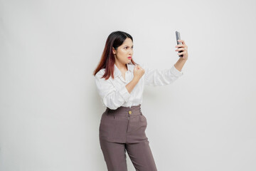 Naklejka premium A dissatisfied young Asian woman looks disgruntled wearing white shirt irritated face expressions holding her phone