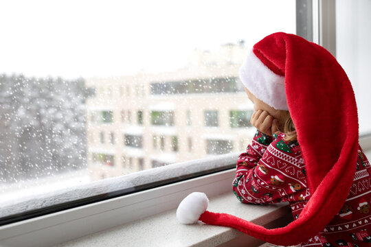 Sad alone child girl in Christmas red Santa hat looks at window and waiting holidays. Mental health