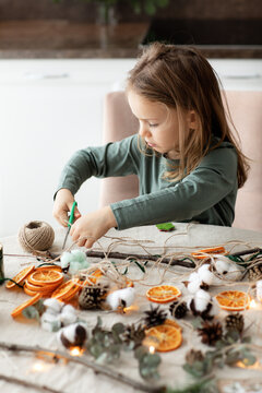 Winter holiday season. child little toddler girl making handmade Christmas garland home decorations from natural ECO materials. Vertical