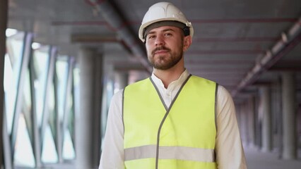 Portrait of successful young architect in hardhat and safety vest looking in camera on blurred background. Bearded male specialist walks on construction site of building with panoramic windows closeup