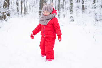 Fototapeta na wymiar Cute little girl in pink snowsuit and knitted hat and scarf plays with snow in winter forest