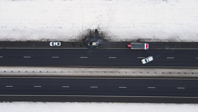 Disaster - burned out car on the edge of the motorway in winter, aerial view. Near the fire and police cars. Help and rescue concept, 911 service. White snow on the side of the road. UHD 4K