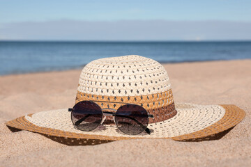 Straw hat with beautiful sunglasses on sand near sea. Beach accessory