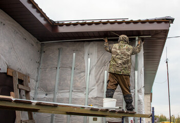 A man installs metal supports on the walls of the house for siding