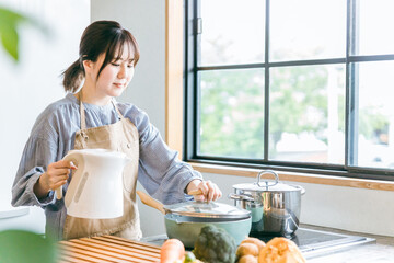 キッチンで電気ケトルのお湯を鍋に入れるエプロン姿のアジア人女性