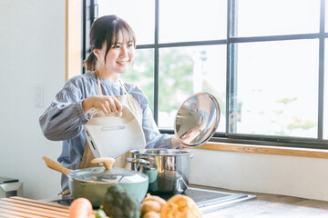 キッチンで電気ケトルのお湯を鍋に入れるエプロン姿のアジア人女性

