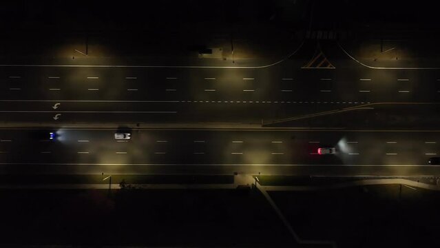 An aerial view over illuminated, parallel roads at night. The camera tilted straight down, truck left along the road, past an intersection with a few cars driving by.