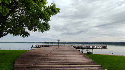 A peaceful atmosphere under the cloudy sky around the lake with wooden dock, meadow and tree viewed
