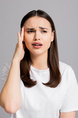 Sad exhausted young girl woman with headache suffering from migraine standing on grey background in studio isolated closing eyes holding arm near head.