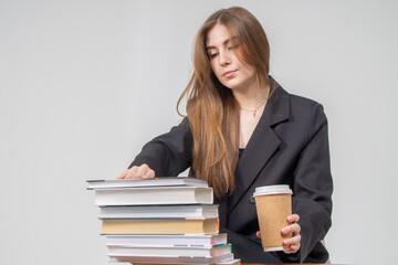 A beautiful young girl sits in front of a stack of books with coffee on a white background. The model is wearing a black jacket and trousers.