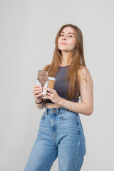 A pretty young brown-haired girl holds a bar of chocolate and a glass of coffee in her hands. The model is wearing a gray top and blue jeans, she smiles sweetly