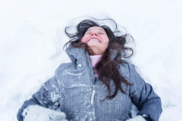 Laughing woman lies on the snow. Pretty brunette enjoys winter. Top view.