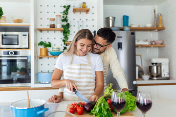 Affectionate young man kissing his wife while she's making breakfast. Beautiful young couple is talking and smiling while cooking healthy food in kitchen at home.Man is kissing his girlfriend in cheek
