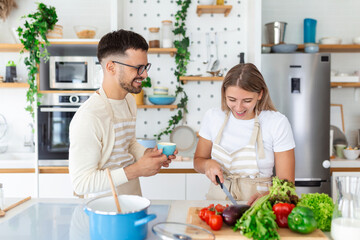 Affectionate young man kissing his wife while she's making diner. Beautiful young couple is talking and smiling while cooking healthy food in kitchen at home.