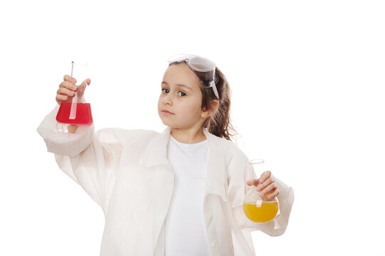 Thoughtful Caucasian little girl, a young scientist chemist in safety goggles and white lab coat, conducts science experiment in chemistry class, isolated on white background with advertising space.