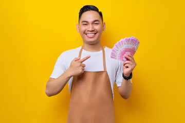 smiling young Asian man barista barman employee wearing brown apron working in coffee shop, pointing a finger at rupiah cash money in hand isolated on yellow background. Small business startup concept